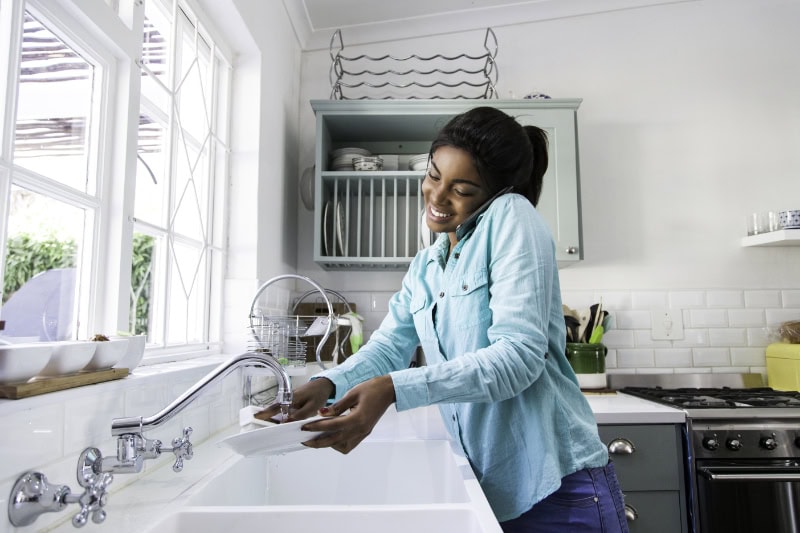 Top 5 Plumbing Upgrades to Include in Your Kitchen Remodel. African female doing the dishes while having a conversation on her mobile phone, laughing. Cape Town, Western Cape, South Africa.