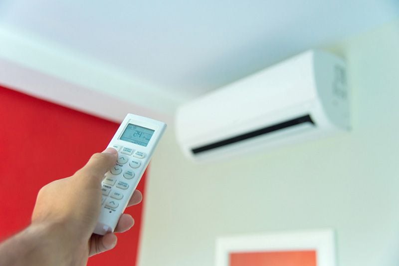 Ductless ACs Improve Indoor Air Quality and Control Humidity. Image shows ductless unit mounted on wall and a hand holding the remote.